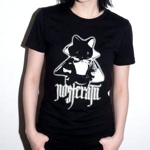 Puede incluir: Una camiseta negra de manga corta con un estampado gráfico blanco de un gato vestido con una capa de cuello alto. Debajo del gato, la palabra "nosferatu" está impresa en una fuente blanca estilizada de inspiración gótica con efecto.