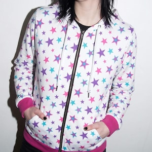 May include: A white zip-up hoodie featuring an all-over print of purple, pink, and turquoise stars. The garment includes a black front zipper, a drawstring hood with white cords, and solid pink ribbed cuffs and a pink ribbed hem at the waist.