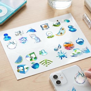 May include: A sheet of various stickers with a glossy finish. The stickers feature a variety of designs, including a cat, a dolphin, musical notes, and a fishbowl. The stickers are in shades of blue, green, and white.