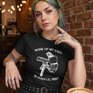 May include: Black t-shirt with the text "NONE OF MY EXES IS OVER LIL PEEP" and a graphic of a gun and a cat. The person in the image is wearing multiple rings and has tattoos on their arms and face.