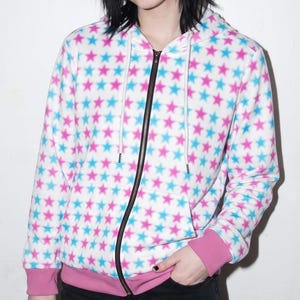 Könnte beinhalten: Ein weißer Kapuzenpullover mit Reißverschluss und einem durchgehenden Muster aus kleinen rosa und hellblauen Sternen. Das Kleidungsstück verfügt über einen schwarzen Frontreißverschluss, eine Kapuze mit weißen Kordeln sowie rosa Rippbündchen.