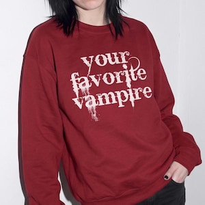 以下が含まれることがあります： 胸元に「your favorite vampire」という白い文字がプリントされた、ダークレッドのクルーネックトレーナー。文字はかすれたような滴るデザインで、長袖で袖口と襟元はリブ編みになっています。