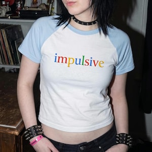 May include: A white and light blue raglan t-shirt with the word impulsive printed in multicolored letters across the chest. The shirt features short sleeves and a crew neckline, paired with black jeans and black spiked wristbands.
