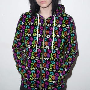 May include: A black zip-up hoodie featuring an all-over repeating pattern of colorful brass knuckles in shades of neon pink, green, orange, purple, blue, and white. The garment includes a hood with white drawstring cords and a central metal zipper.