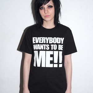 Puede incluir: Una camiseta negra de manga corta con el texto impreso en blanco "EVERYBODY WANTS TO BE ME!!" en el pecho. La prenda tiene cuello redondo y un corte est&aacute;ndar, confeccionada en tela negra lisa con letras blancas llamativas.