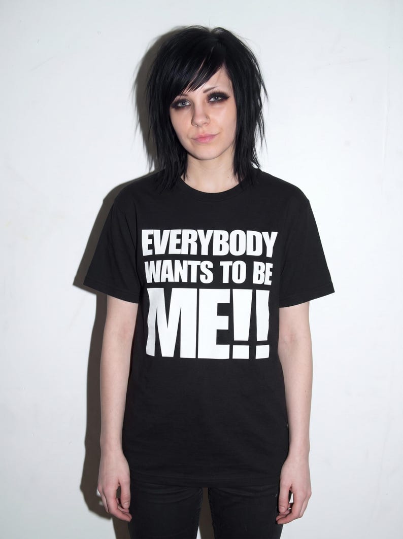 Puede incluir: Una camiseta negra de manga corta con el texto impreso en blanco "EVERYBODY WANTS TO BE ME!!" en el pecho. La prenda tiene cuello redondo y un corte est&aacute;ndar, confeccionada en tela negra lisa con letras blancas llamativas.