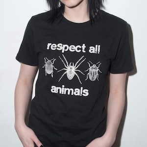 May include: A black short-sleeved t-shirt featuring a white graphic print of a spider flanked by two beetles. The text "respect all" is printed above the insects, and the word "animals" is printed below them in a bold, rounded white font.