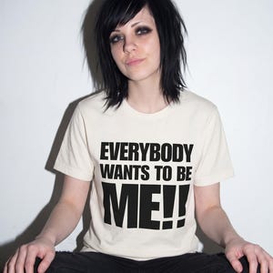 Puede incluir: Una camiseta de manga corta de color crema con el texto negro "EVERYBODY WANTS TO BE ME!!" impreso en una fuente negrita sin remates en el pecho. La prenda tiene un cuello redondo cl&aacute;sico y un dobladillo recto.