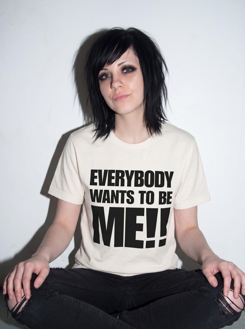 Puede incluir: Una camiseta de manga corta de color crema con el texto negro "EVERYBODY WANTS TO BE ME!!" impreso en una fuente negrita sin remates en el pecho. La prenda tiene un cuello redondo cl&aacute;sico y un dobladillo recto.