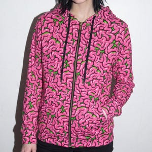 May include: A zip-up hooded sweatshirt featuring an all-over print of pink brain-like structures with green dripping accents. The garment includes a full-length metal zip, long sleeves, two front pockets, and black drawstring cords at the hood.