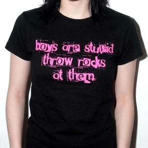 Puede incluir: Una camiseta negra de manga corta con el texto "boys are stupid throw rocks at them" impreso en un estilo desgastado y color rosa brillante en el pecho. La prenda tiene cuello redondo y un corte estándar.