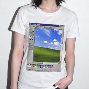 May include: A white short-sleeved t-shirt featuring a graphic print of the classic Windows Paint software interface. The screen displays a green grassy hill under a blue sky with white clouds, surrounded by toolbars, menus, and a color palette.