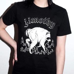 Puede incluir: Una camiseta negra de manga corta con un gráfico blanco de un mapache caminando entre llamas. Sobre el mapache, el texto "Jimothy" está impreso en una fuente blanca estilizada en la parte frontal de la prenda de vestir.