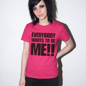 Puede incluir: Una camiseta de manga corta de color rosa brillante con el texto "EVERYBODY WANTS TO BE ME!!" impreso en letras may&uacute;sculas negras y negritas en el pecho. La prenda tiene un cuello redondo cl&aacute;sico y un dobladillo recto, de corte sencillo.