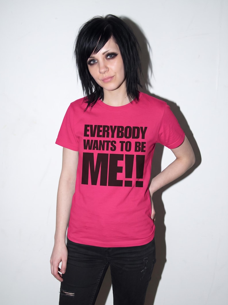 Puede incluir: Una camiseta de manga corta de color rosa brillante con el texto "EVERYBODY WANTS TO BE ME!!" impreso en letras may&uacute;sculas negras y negritas en el pecho. La prenda tiene un cuello redondo cl&aacute;sico y un dobladillo recto, de corte sencillo.