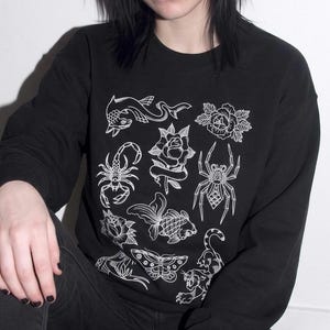 May include: A black long-sleeve sweatshirt featuring a white graphic print of various tattoo-style illustrations, including a scorpion, a spider, a fish, a moth, a panther, and several roses arranged in a grid pattern on the front.
