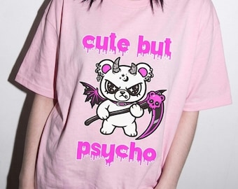 May include: A light pink short-sleeve t-shirt featuring a graphic of a white bear with horns, bat wings, and a scythe. The text "cute but" appears above the bear and "psycho" appears below, both in a dripping pink font style.
