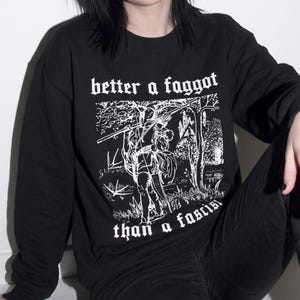 May include: A black long-sleeve sweatshirt featuring a white graphic print of a figure carrying a scythe in a wooded landscape. The text "better a faggot than a fascist" is printed in a gothic-style font above and below the illustration.
