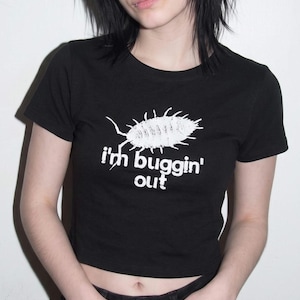 May include: A black short-sleeved crop top featuring a white graphic of an isopod insect centered on the chest. Below the insect, the text "i'm buggin' out" is printed in a white, rounded sans-serif font. The garment has a crew neckline.