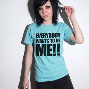 Puede incluir: Una camiseta de manga corta de color azul claro con el texto negro "EVERYBODY WANTS TO BE ME!!" impreso en una fuente negrita sin remates en el pecho. La prenda tiene cuello redondo y un corte est&aacute;ndar de tela lisa.