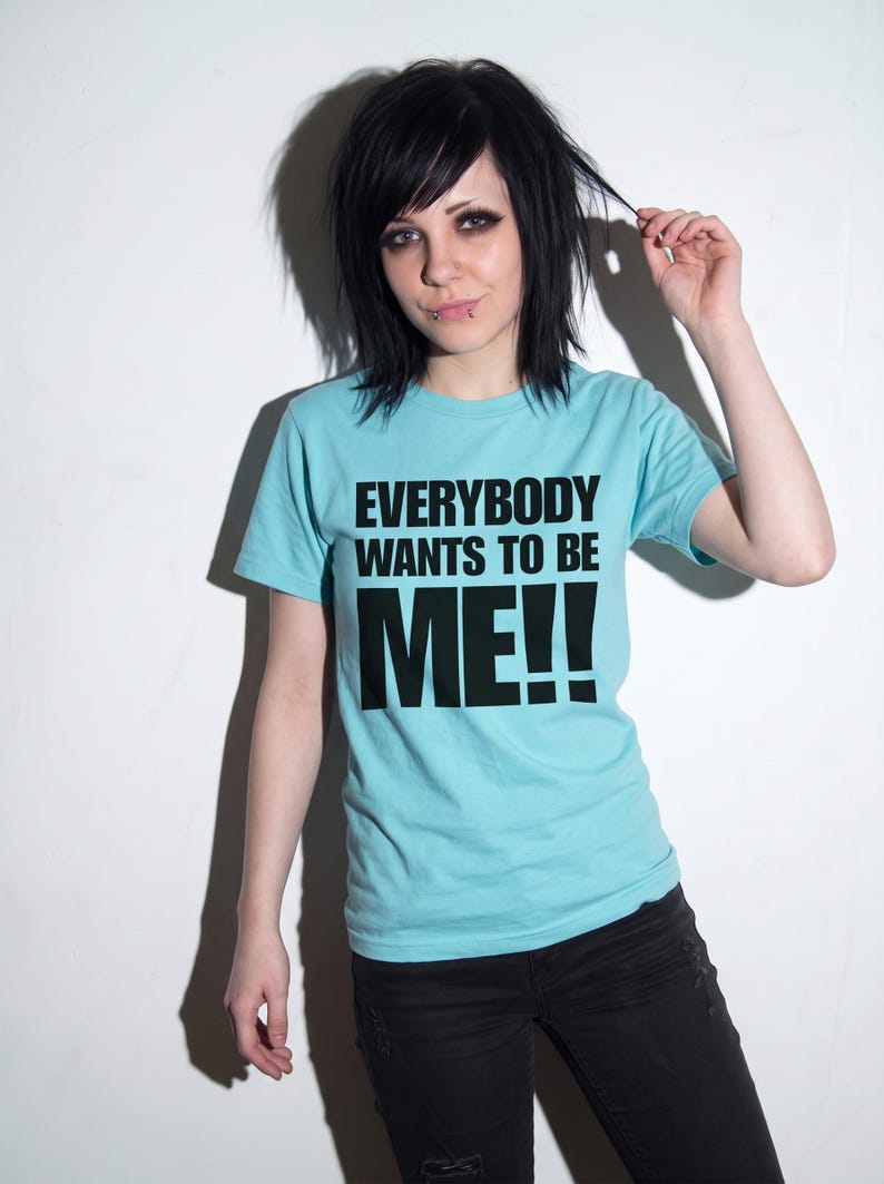 Puede incluir: Una camiseta de manga corta de color azul claro con el texto negro "EVERYBODY WANTS TO BE ME!!" impreso en una fuente negrita sin remates en el pecho. La prenda tiene cuello redondo y un corte est&aacute;ndar de tela lisa.