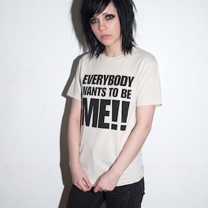 Puede incluir: Una camiseta de manga corta de color crema con el texto negro "EVERYBODY WANTS TO BE ME!!" impreso en la parte delantera. La camiseta tiene cuello redondo y se combina con unos vaqueros negros desgastados, creando un estilo sencillo.