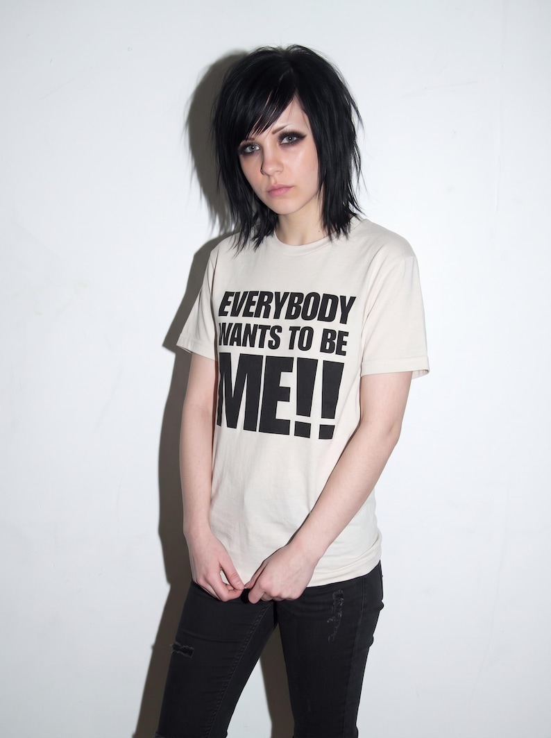 Puede incluir: Una camiseta de manga corta de color crema con el texto negro "EVERYBODY WANTS TO BE ME!!" impreso en la parte delantera. La camiseta tiene cuello redondo y se combina con unos vaqueros negros desgastados, creando un estilo sencillo.