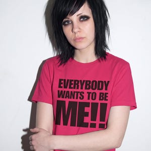 Puede incluir: Una camiseta de manga corta de color rosa brillante con el texto negro "EVERYBODY WANTS TO BE ME!!" impreso en una fuente negrita sin remates en el pecho. La prenda tiene un cuello redondo cl&aacute;sico y un dobladillo recto.