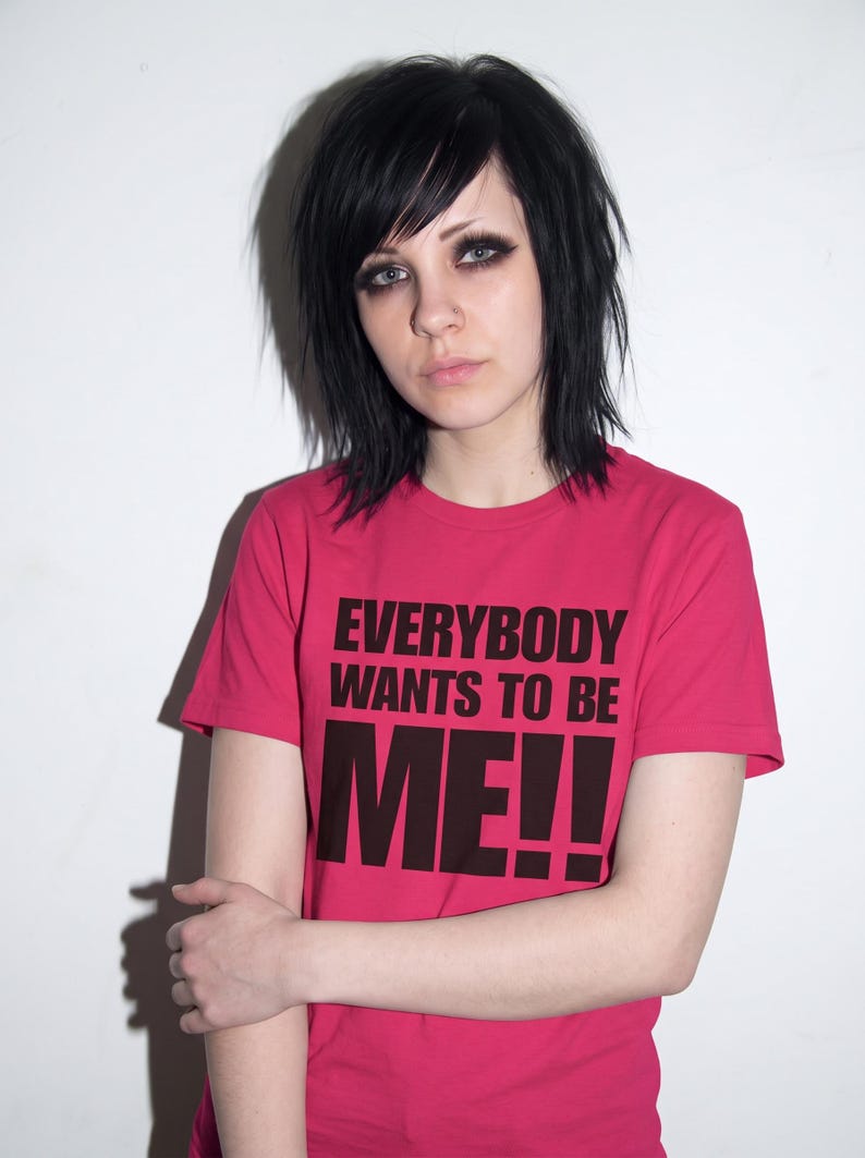 Puede incluir: Una camiseta de manga corta de color rosa brillante con el texto negro "EVERYBODY WANTS TO BE ME!!" impreso en una fuente negrita sin remates en el pecho. La prenda tiene un cuello redondo cl&aacute;sico y un dobladillo recto.