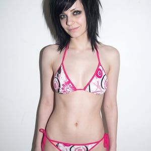 K&ouml;nnte beinhalten: Ein zweiteiliger Bikini mit wei&szlig;em Grund und einem grafischen Muster aus rosa Blumen und schwarzen Kreisumrissen. Das Oberteil hat Triangel-Cups mit rosa Einfassung und d&uuml;nnen Tr&auml;gern, w&auml;hrend das Unterteil seitliche Bindeb&auml;nder besitzt.