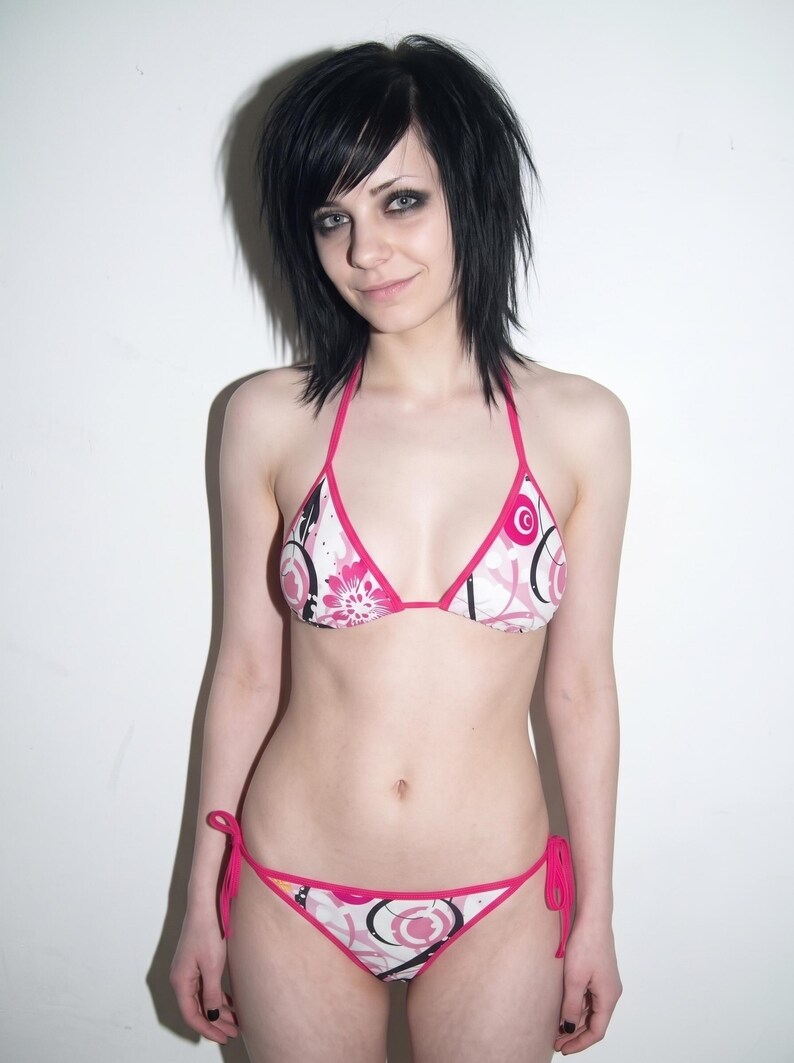 K&ouml;nnte beinhalten: Ein zweiteiliger Bikini mit wei&szlig;em Grund und einem grafischen Muster aus rosa Blumen und schwarzen Kreisumrissen. Das Oberteil hat Triangel-Cups mit rosa Einfassung und d&uuml;nnen Tr&auml;gern, w&auml;hrend das Unterteil seitliche Bindeb&auml;nder besitzt.