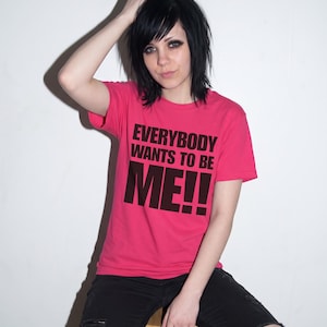Puede incluir: Una camiseta de manga corta de color rosa brillante con el texto negro en negrita "EVERYBODY WANTS TO BE ME!!" impreso en el pecho. La prenda se combina con unos vaqueros negros desgastados con roturas visibles en las rodillas.