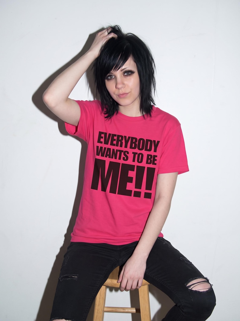 Puede incluir: Una camiseta de manga corta de color rosa brillante con el texto negro en negrita "EVERYBODY WANTS TO BE ME!!" impreso en el pecho. La prenda se combina con unos vaqueros negros desgastados con roturas visibles en las rodillas.