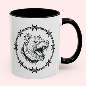 Può includere: Una tazza in ceramica bianca con manico e bordo neri, caratterizzata da un'illustrazione a linee nere di un cane ringhiante con collare a punte, racchiusa in una cornice circolare di filo spinato centrata sulla parte anteriore.