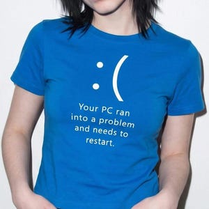 May include: A bright blue short-sleeved t-shirt featuring a white graphic of a sad emoticon and the text: Your PC ran into a problem and needs to restart. The design mimics a computer system error screen printed on the front of the garment.
