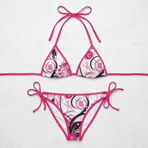 K&ouml;nnte beinhalten: Ein zweiteiliger Bikini mit wei&szlig;em Hintergrund und einem abstrakten Blumen- und Wirbelmuster in leuchtendem Pink und Schwarz. Das Oberteil hat Triangel-Cups mit pinkfarbenen B&auml;ndern, das Unterteil verf&uuml;gt &uuml;ber seitliche Bindeb&auml;nder.