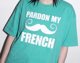May include: A short-sleeved teal t-shirt featuring the white text "PARDON MY" above a large white graphic of a handlebar mustache, with the word "FRENCH" printed in white capital letters underneath the mustache design.