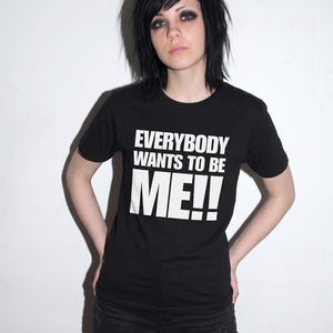 Puede incluir: Una camiseta negra de manga corta con el texto blanco "EVERYBODY WANTS TO BE ME!!" impreso en una fuente negrita y sin remates en el pecho. La prenda tiene un cuello redondo est&aacute;ndar y un dobladillo recto.