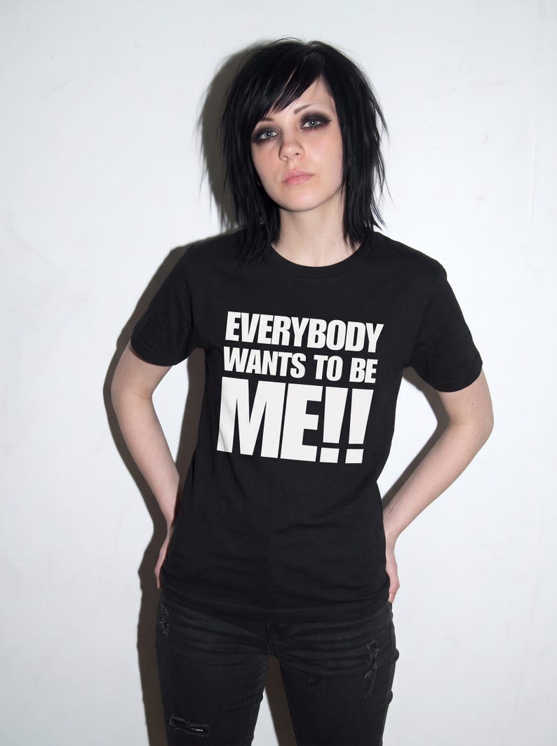 Puede incluir: Una camiseta negra de manga corta con el texto blanco "EVERYBODY WANTS TO BE ME!!" impreso en una fuente negrita y sin remates en el pecho. La prenda tiene un cuello redondo est&aacute;ndar y un dobladillo recto.
