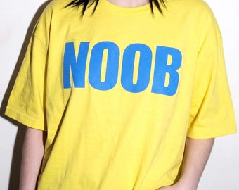 May include: A bright yellow short-sleeved t-shirt featuring the word NOOB printed in large, bold, blue capital letters across the chest. The garment has a classic crew neckline and a relaxed fit, providing a simple and casual appearance.