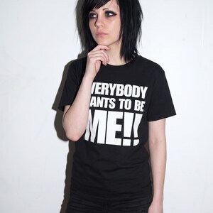 Puede incluir: Una camiseta negra de manga corta con letras blancas en bloque que dicen EVERYBODY WANTS TO BE ME!!. La prenda tiene cuello redondo y est&aacute; confeccionada en tela negra lisa. El texto est&aacute; impreso en una fuente negrita sobre el pecho.