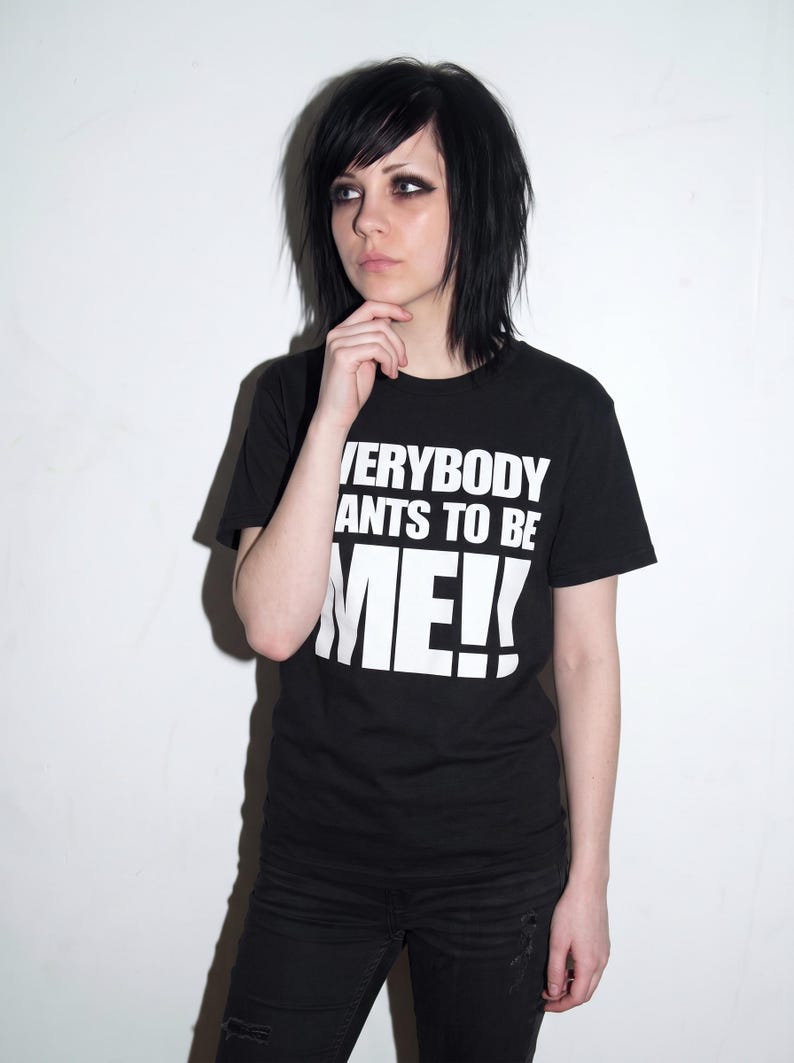 Puede incluir: Una camiseta negra de manga corta con letras blancas en bloque que dicen EVERYBODY WANTS TO BE ME!!. La prenda tiene cuello redondo y est&aacute; confeccionada en tela negra lisa. El texto est&aacute; impreso en una fuente negrita sobre el pecho.