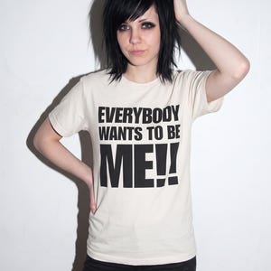Puede incluir: Una camiseta de manga corta de color crema con el texto negro "EVERYBODY WANTS TO BE ME!!" impreso en una fuente negrita sin remates en el pecho. La camiseta tiene un cuello redondo cl&aacute;sico y un dobladillo recto.