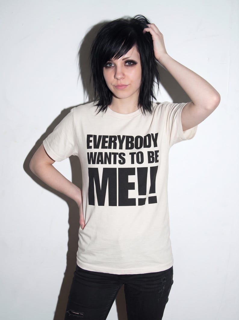 Puede incluir: Una camiseta de manga corta de color crema con el texto negro "EVERYBODY WANTS TO BE ME!!" impreso en una fuente negrita sin remates en el pecho. La camiseta tiene un cuello redondo cl&aacute;sico y un dobladillo recto.