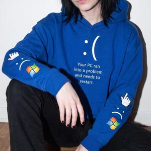 May include: A bright blue hooded sweatshirt featuring a white sad face emoticon and the text "Your PC ran into a problem and needs to restart." The sleeves display a white pixelated cursor icon and a colorful four-pane window logo graphic.