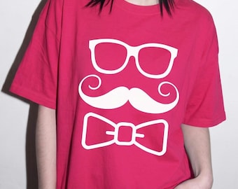 May include: A bright pink short-sleeved t-shirt featuring a large white graphic print on the front. The design consists of a pair of thick-rimmed glasses positioned above a stylized handlebar mustache and a classic bow tie centered below.