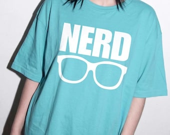May include: A turquoise short-sleeved t-shirt featuring the word NERD printed in large white capital letters above a graphic of white thick-rimmed eyeglasses. The shirt has a crew neckline and a relaxed fit, made of a solid-colored cotton fabric.