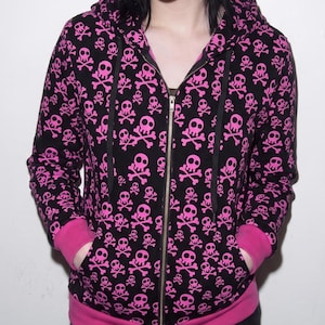 Puede incluir: Una sudadera negra con cremallera y capucha que presenta un patrón repetitivo de calaveras y tibias cruzadas en color rosa brillante. La prenda incluye una capucha con cordones negros, cremallera frontal completa, dos bolsillos y puños y dobladillo acanalados en rosa sólido.