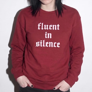 May include: A maroon long-sleeve crewneck sweatshirt featuring the white text "fluent in silence" printed in a gothic-style font across the chest. The garment has ribbed cuffs and a ribbed hem, providing a standard fit for casual wear.