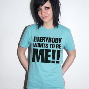 Puede incluir: Una camiseta de manga corta de color azul claro con el texto negro "EVERYBODY WANTS TO BE ME!!" impreso en una fuente negrita sin remates en el pecho. La prenda tiene un cuello redondo cl&aacute;sico y un dobladillo recto.